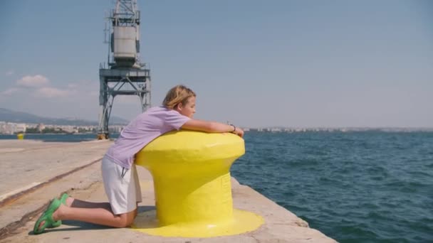 紫色のTシャツと白いショートパンツの男の子は 背景に大きなクレーンで海を見ながら黄色い港のボラードを走ります — ストック動画