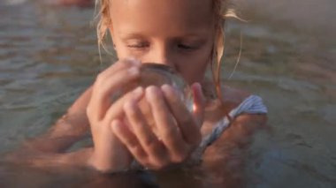Bir kız, kısmen deniz suyuna batmış, elinde cam bir top tutuyor, dikkatle içine bakıyor, yüzünde düşünceli bir ifadeyle.