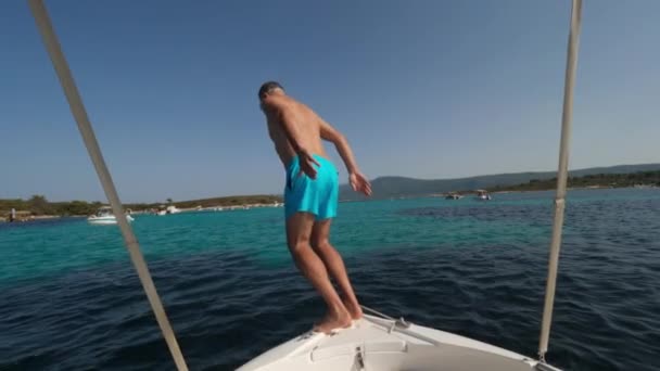 一名身穿蓝色短裤的男子从一艘白色汽船的船头跳入大海 在沿海风景和其他游艇的衬托下 — 图库视频影像