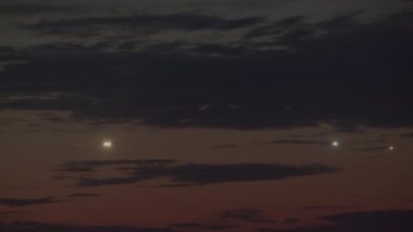 Alacakaranlık gökyüzünde, farklı uzaklıklarda yer alan üç set uçak ışığı göze çarpıyor. Ufukta koyu bulutlar ve kırmızımsı renkler var.