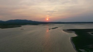 Sakin bir günbatımının havadan görünüşü engin bir gölün yumuşak dalgaları üzerine sıcak bir parıltı saçıyor, yemyeşil arazi ve karanlık bir gökyüzünün altındaki uzak tepelerle çevrili.