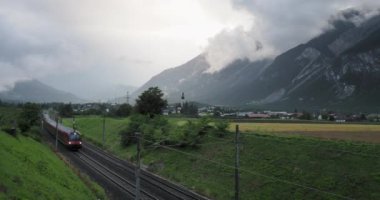 Bir ekspres tren, bulutlu bir dağ zeminiyle kırsal alanda hızla ilerlerken Avusturya Alpleri 'nde kilise kulesi olan bir köyü geçiyor.