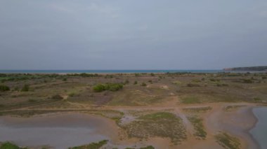 Uçurumlarla çevrili uzak mavi denize doğru giden yol ve su cepleri bulunan pastel bir gökyüzünün altındaki geniş kıyı çalılıklarının hava görüntüsü.