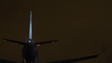 İniş ışıkları açık, gece gökyüzünün altında, parlak pist ışıklarıyla çevrili bir uçak.