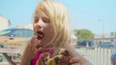 Pembe tişörtlü sarışın bir kız sokakta kuru üzümlü çörek yiyor bulanık bir şehrin arka planında