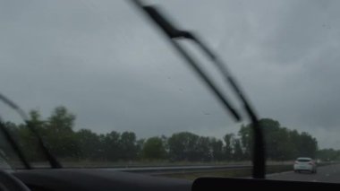 Yağmurla ıslanmış bir otoyolda araba sürerken cam silecekleri açık bir şekilde ön camdaki damlacıklara ve dışarıdaki yol ve ağaçların bulanık görüntüsüne odaklanıyor.