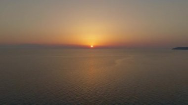 İnsansız hava aracı geri çekiliyor, sıcak gölgeler içinde sakin denizin üzerinde gün batımını yakalıyor.
