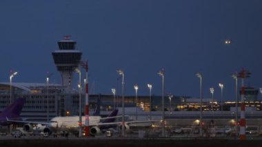 Akşam havaalanı ve yaklaşan bir uçak. Kontrol kulesi aydınlandı ve çeşitli parkedilmiş uçaklar aydınlatma terminalinin arka planında görülebiliyor.