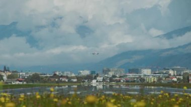 Bir uçak, bir çiçek tarlası boyunca görünen, dağlardan oluşan bir zemin ve bulutlu gökyüzü ile bir şehrin üzerine inmek için yaklaşıyor. Avusturya Alpleri. Innsbruck Havaalanı