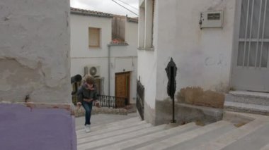 Gri ceketli bir ağabey, kız kardeşini beyaz evlerin arasında beton merdivenlerden yukarı taşıyor.