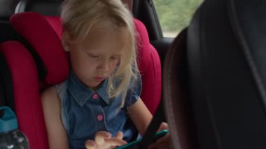 Genç bir kız, parlak kırmızı bir araba koltuğunda otururken tablete sarılmış vaziyette, aracın içi ve su şişesi de görüş alanımızda.