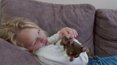 Kanepede uzanmış genç bir kız Chihuahua köpeğini nazikçe kucaklıyor.