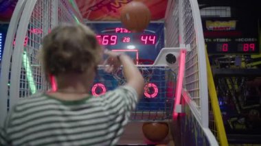 Çizgili tişörtlü bir çocuk atari salonu basketbolu oynuyor, topu dijital bir skorbordun altında potaya atıyor ve arka planda zamanı gösteriyor ve sayı yapıyor.