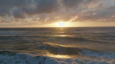 İnsansız hava aracı, gün batımında dalgaların yavaş çekim videosunu çekiyor. Güneş ışığı bulutlu bir gökyüzünün altında suya yayılıyor.