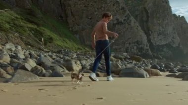 Kot pantolon ve süveter giyen genç bir çocuk kumlu bir sahilde büyük kayalar ve arka planda bir uçurum olan bir Chihuahua yürür.