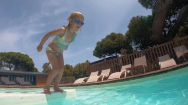 Mayo giymiş ve gözlüklü genç bir kız su altında yüzüyor. Etrafı berrak turkuaz suyla çevrili. Yavaş çekim.