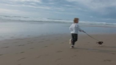 Genç bir kız, dalgaların arka planında kumlu bir sahil boyunca bir chihuahua ile güçlü bir şekilde koşuyor.