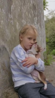 Sarı saçlı, çizgili kazaklı genç bir kız duvara yaslanmış, kahverengi ve beyaz bir Chihuahua ile etkileşimde ve dışarıdaki kameraya bakıyor.