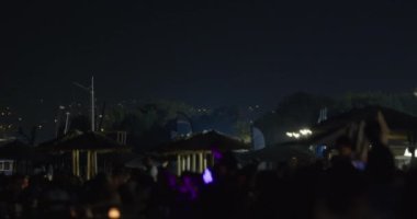 Sahilde, şemsiyelerle aydınlatılmış insan siluetleri, uzak şehir ışıkları ve arka planda yükselen bir uçağın belirgin ana hatlarıyla dolu canlı bir gece sahnesi.