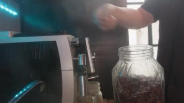Kadın kahve makinesini kavanozdan dolduruyor. Mutfakta buhar yükseliyor.