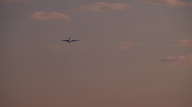 Pembe ve mor tonlarıyla alacakaranlık gökyüzünün arka planında uçan bir uçak.