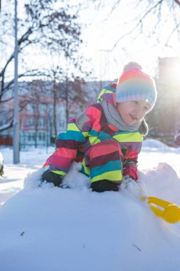 Renkli bir kar elbisesi içindeki çocuk kış boyunca bir park alanında kar yığınına tırmanıyor. Arka planda binalar ve ağaçlar görünür.