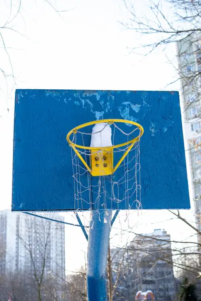 Gündüz vakti kar yağan bir basketbol potası, mavi bir arka tahtası ve sarı kenarlı.