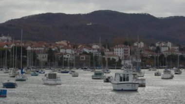 SAN SEBASTIAN, SPAIN - HAZİRAN 03, 2024: Bulutlu bir gökyüzünün altındaki yamaçta yuva yapmış bir köyün önündeki yelkenli ve motorlu tekne dolu bir yat limanı