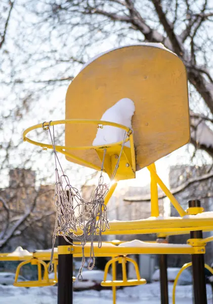 Yırtık bir ağ ile sarı basketbol potası boş bir oyun parkında karla kaplı, kış boyunca açık alanların durgunluğunu vurguluyor.