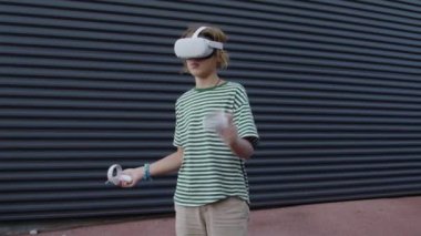 Çizgili tişört giyen ve virajlı bir duvara yaslanmış, VR kulaklıklı ve joystickli bir genç açık havada sanal gerçeklik oyunundan hoşlanıyor.