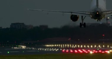 Uçak alacakaranlıkta ıslak bir piste iniyor. İniş ışıkları pisti aydınlatıyor.