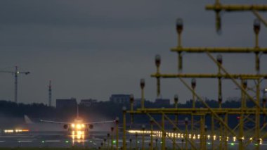 Düşük ışık koşullarında, sarı pist ışıkları ve gri gökyüzü olan ıslak bir pistte kalkış yapan yolcu uçağı, arka planda vinçler görülüyor.