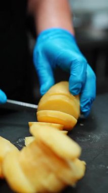 Profesyonel şef mavi eldiven giyip patatesi dikkatlice siyah kesim tahtasına ince parçalar halinde dilimliyor. Mutfak tekniği ve profesyonel yemek hazırlama becerileri sergiliyor.