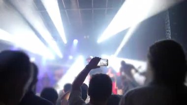 Konsere gidenler canlı müzik performansı yakalıyorlar, ellerinde akıllı telefonlar ve parlak sahne ışıkları altında çalan bando, modern müzik deneyimini ve dijital paylaşım kültürünü sergiliyorlar.