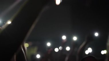 Konser izleyicileri performansı kaydediyor, karanlık mekanı akıllı telefon ışıklarıyla aydınlatıyor, kalp şeklindeki objeyi sallıyor, canlı müzik etkinliği sırasında toplu tutku ve enerjiyi ifade ediyor