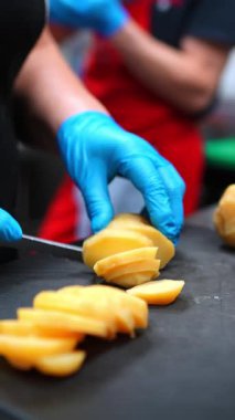 Profesyonel aşçı eldivenli eller patatesi üniforma içinde ince dilimler siyah kesme tahtasında, aşçılık tekniğini ve yemek hazırlama uzmanlığını vurguluyor.