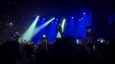 Canlı müzik etkinliğindeki izleyiciler, performansı yakalamak için ellerinde akıllı telefonlar, dinamik ışıklandırma ve enerjik ambiyansla çevrili.