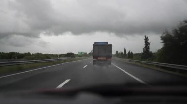 Bir kamyon yeşil ve ağaçlarla çevrili bir otoyolda ilerliyor, bulutlu gökyüzü ve yağmur karamsar bir atmosfer yaratıyor.