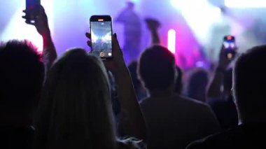 Karanlık bir kalabalığın içindeki insanlar, havada duran akıllı telefonlar ile bir konseri filme çekiyorlar. Arkadan çekiyorlar ve yüzleri gözükmüyor.