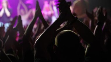 Seyirciler, renkli sahne ışıkları ve canlı bir atmosferle aydınlatılan canlı konser sırasında sevinçle el kaldırıyorlar.