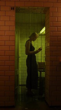 Koyu renk bar tuvaletinde sarı yeşil ışığın altında zincir perdenin arkasında duran kadın.