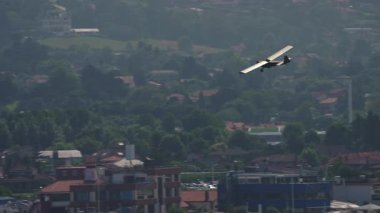 Ahşap gövdeli eski model eğitim uçağı açık kokpitli ve cam kaplamasız şehir binalarının üzerinde uçuyor.