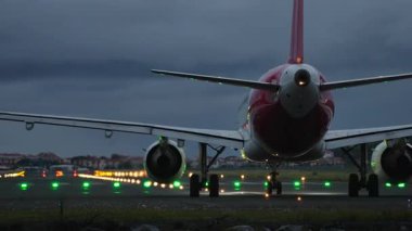 Uçak gece podyumda, aydınlık ışıklar ve çarpıcı bulutlu gökyüzü ile çevrili, havacılık ve seyahat atmosferi sergileniyor.