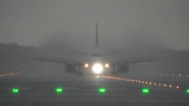 Uçak yağmurlu ve sisli koşullarda piste yaklaşır, aydınlatılmış ışıklar inişe rehberlik eder, zorlu hava koşullarında havacılığı gösterir