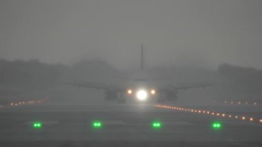 Yoğun sis altında piste yaklaşan uçak, yoğun yağmur sırasında yolu aydınlatan parlak ışıklarla aydınlanıyor.