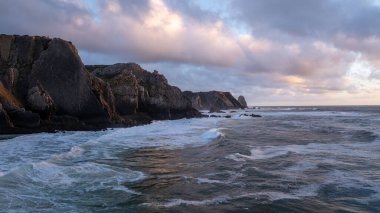 Güneş, Portekiz 'in Sintra kentindeki Praia da Adraga sahilindeki dalgaların üzerinde batıyor ve alacakaranlık gökyüzünün altında dramatik bir deniz manzarası yaratıyor.