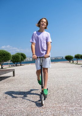 Kaygısız genç çocuk, deniz kenarındaki canlı bir parkta scooter 'a biniyor, güneşleniyor ve keyifli bir yaz gününün tadını çıkarıyor.