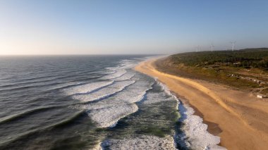 Portekiz, Nazare 'nin kumlu plajlarında dalgaların uçuşması, yamaçlarda temiz enerji üreten rüzgar türbinleri.