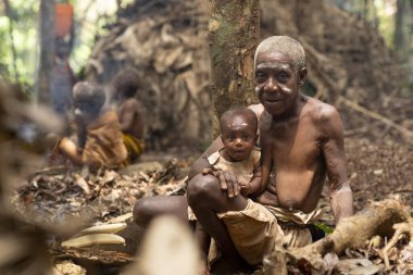 Yaşlı bir pigme kadın, Kamerun 'un tropikal ormanında kucağında küçük bir çocuk tutuyor. Yüzleri geleneksel boyalar ile süslenir ve arkalarında, günlük yaşam yaprakların oluşturduğu sığınakların etrafında gelişir. İçler acısı anı yakalıyor.