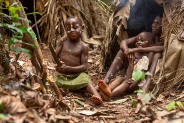 Bir pigme adam, Kamerun ormanının ortasında, geleneksel bir yaprak kulübesinin önünde iki çocukla oturuyor. Bir çocuk palayla oynarken diğeri büyükler ona sarılıyor..